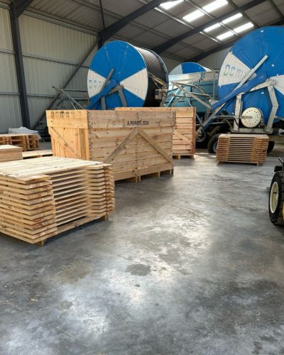 Entrepôt avec bobines bleues de câbles, caisses "A PRIORI", et planches en bois au sol en béton.