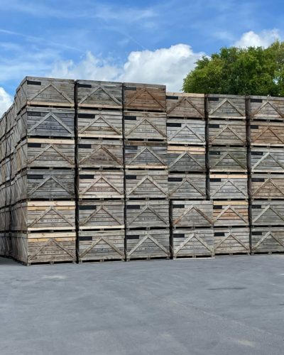 Empilement de grandes caisses en bois, sol en béton, ciel dégagé, arbres en arrière-plan.