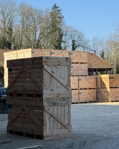 Empilement de grandes caisses en bois sur gravier entouré d'arbres dénudés.